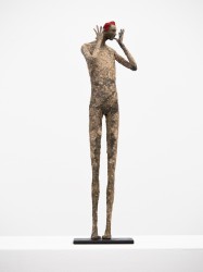 An elongated figure with bright red hair gestures to the viewer in this expressive paper mache sculpture by Paul Duval.